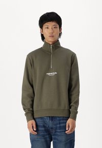 Maslinasto zeleni zip-up dukserica s visokim ovratnikom, s bijelim logotipom na prsima, izrađena od mekog materijala s opuštenim krojem.