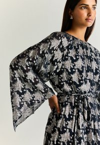 Abito lungo e fluente in tessuto navy trasparente con motivi floreali argentati. Presenta ampie maniche a campana e una vita cintrata per una silhouette definita.