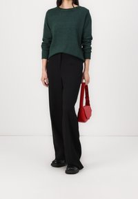 Personne portant un pull vert foncé, un pantalon large noir, des chaussures noires, tenant un petit sac à main rouge sur fond blanc.