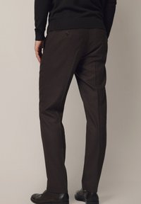 Brązowe spodnie od garnituru o prostym kroju, wykonane z gładkiego materiału, z tylną kieszenią zapinaną na guzik. W zestawie z czarnymi butami.
