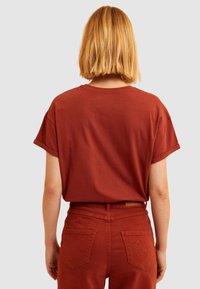 Camiseta de algodón de manga corta en color óxido, vista desde atrás. Combinada con pantalones de talle alto a juego que presentan detalles de bolsillos.