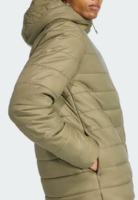 Homme portant une veste matelassée chaude de couleur vert olive avec une capuche, se tenant de côté et reposant sa main dans la poche de la veste.