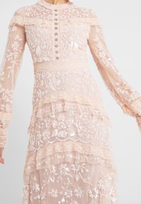 Ljust rosa, långärmad klänning med skiktade volanger, intrikat blommönsterbroderi, paljetter och dekorativa knappar nedanför.