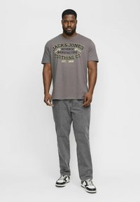 Gray t-shirt with 'JACK & JONES' logo in yellow and black, paired with gray jeans and black-and-white sneakers. Casual fit.
