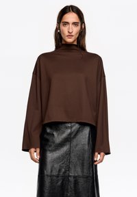 Brązowa, krótka bluzka z długim rękawem wykonana z gładkiej tkaniny, zestawiona z błyszczącą czarną skórzaną spódnicą o prostej sylwetce i długości do kolan.