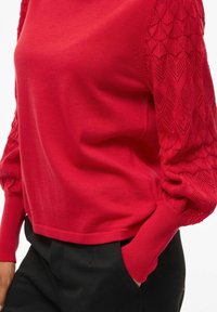 Pull rouge avec un corps uni et des manches texturées à motif en losange. Le design comprend des poignets ajustés et un ourlet court.