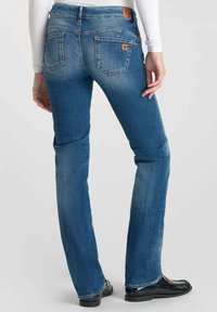 Jean en denim bleu avec une coupe ajustée et une légère évasée, présentant un patch en cuir marron et deux poches arrière avec des détails de couture.