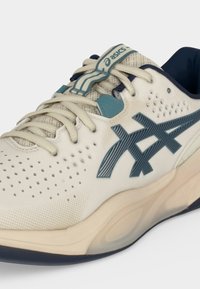 Λευκό και μπλε παπούτσι Asics με διάτρητο επάνω μέρος, κορδόνια, επενδυμένη σόλα και λογότυπο της μάρκας στο πλάι και στη γλώσσα.