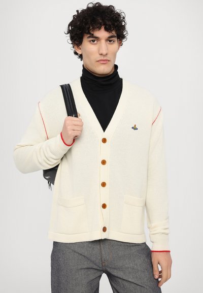 Creme cardigan med træknapper, røde accenter på ærmerne, V-halsdesign og forlommer. Båret over en sort turtleneck.