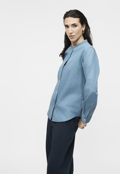 Γυναίκα με μακριά σκούρα μαλλιά που φορά χρυσούς κρίκους στα αυτιά, ανοιχτό μπλε πουκάμισο με κουμπιά και σκούρο παντελόνι, που στέκεται μπροστά σε απλό λευκό φόντο.