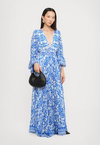 Abito maxi blu con motivi floreali bianchi, scollatura a V profonda, maniche lunghe a campana e una silhouette fluida. Abbinato a una borsa a mano intrecciata nera.