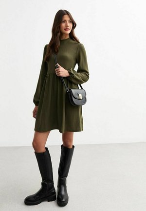 Olijfgroen geribbeld jurk met lange mouwen en elastische taille, gecombineerd met kniehoge zwarte leren laarzen en een zwarte crossover tas.