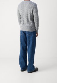 Grauer Kabelstrickpullover mit gerippten Bündchen und Saum, kombiniert mit lockeren blauen Denim-Jeans und schwarzen Schuhen, von hinten betrachtet.