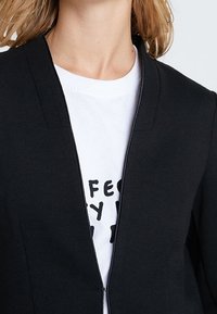 Czarna marynarka o gładkiej fakturze, z otwartym przodem i dekoltem w kształcie litery V, nałożona na białą koszulkę z grafiką i czarnym tekstem.