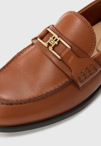 Brown leather loafer with gold metal buckle detail and black sole, featuring detailed stitching on upper and strap.