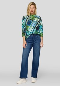 Frau mit blondem Haar trägt ein grün-blau gemustertes Oberteil, blaue gerade geschnittene Jeans und weiße Turnschuhe, steht vor einem einfarbigen Hintergrund.