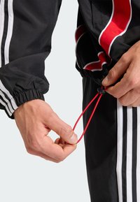 Čierna tepláková súprava s bielymi pruhmi a červenými akcentmi; nastaviteľný šnúrka v ruke; manžety sú elastické; ľahký materiál.