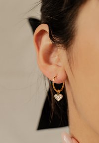Orecchino a cerchio dorato con un ciondolo a forma di cuore argentato, impreziosito da gemme trasparenti, appeso al lobo, che mostra una superficie lucida e un design delicato.