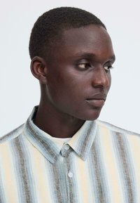 Gestreept overhemd met lichte blauwtinten, geel en grijs. Overhemd met kraag, structuurstof en twee knopen aan de voorkant.