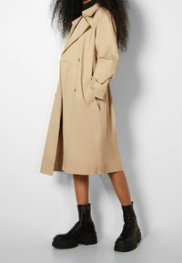Manteau trench beige avec un design croisé, coupe oversize et manches retroussées, associé à des bottines noires à semelles épaisses.