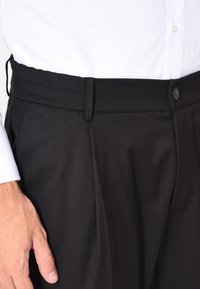 Pantaloni neri con una texture liscia, caratterizzati da una vestibilità su misura, tasche laterali e una chiusura con bottone in vita. Indossati con una camicia bianca.