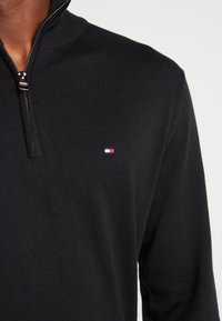 Pull dai collo zip nero realizzato in tessuto a coste. Presenta un piccolo logo con accenti rossi, bianchi e blu sul petto.