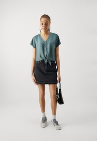 Lys teal kortermet bluse med knyting foran, kombinert med et svart denim miniskjørt. Tilbehør inkluderer en liten svart håndveske og grå sneakers.