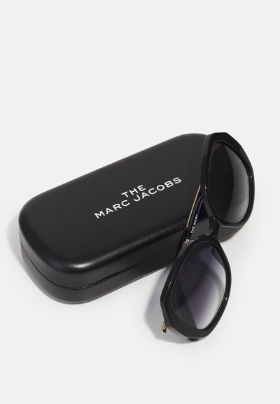 Schwarze Sonnenbrille mit einem achteckigen Rahmen, dunklen, verlaufenden Gläsern und goldenen Akzenten, neben einem schwarzen Lederetui mit weißer Markenbeschriftung platziert.