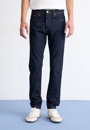 Homme portant un jean en denim bleu foncé et des baskets blanches, debout sur un sol bleu clair devant un fond blanc.