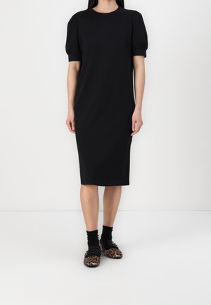 Femme portant une robe noire longueur genou avec des manches courtes bouffantes et des chaussettes noires avec des ballerines à imprimé léopard, debout sur un sol blanc.
