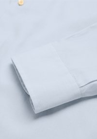 Puño de camisa a rayas de color azul claro, con una textura suave, que presenta un cierre de botón y una costura ordenada a lo largo del borde.