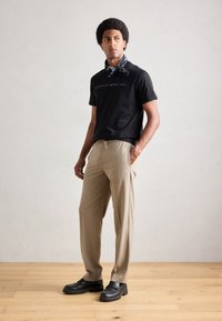 Tricou negru din bumbac cu text imprimat, pantaloni bej cu talie cu șnur și pantofi negri. Modelul poartă o eșarfă modelată în nuanță de bleumarin.