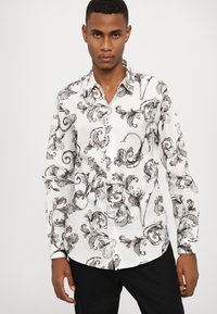 Camisa blanca con patrones florales negros, cuello abotonado y mangas largas. Presenta intrincados remolinos y detalles en texto a lo largo de toda la prenda.