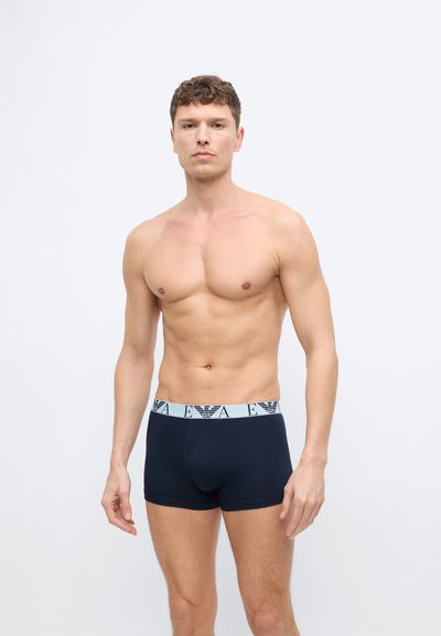 Νεαρός άνδρας με καλή φυσική κατάσταση στέκεται όρθιος, φορώντας μαύρα κοντά εσώρουχα με λευκή ζώνη μέσης που φέρει το λογότυπο EA, σε απλό λευκό φόντο.