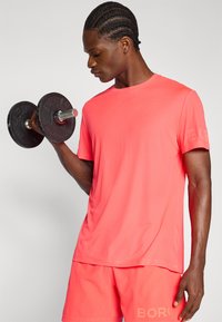 Coral athletic t-shirt with short sleeves, featuring a textured pattern. Model holds a black dumbbell, wearing matching shorts.