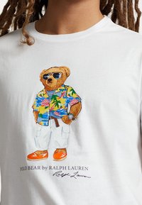 Wit T-shirt met een afbeelding van een beer in een kleurrijk bloemenoverhemd, cargoshort, oranje schoenen en een zonnebril. Onder de afbeelding staat de tekst "POLO BEAR by RALPH LAUREN."
