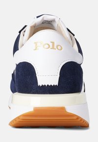 Marinblå mockasneaker med vita läderdetaljer, prydd med en guldig "Polo"-logotyp, vadderad vit sula och en orange gummisula.