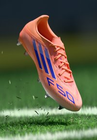 Jasne oranžové futbalové kopačky s modrými pruhmi a nápisom "F50", s textúrovaným povrchom a priehľadnými plastovými špicami, plávajúce nad trávou.