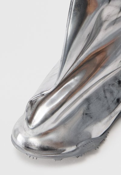 Bottine métallique argentée avec un design froncé, texture lisse et semelle en caoutchouc dentelée. Accent sur une silhouette élégante et contemporaine.