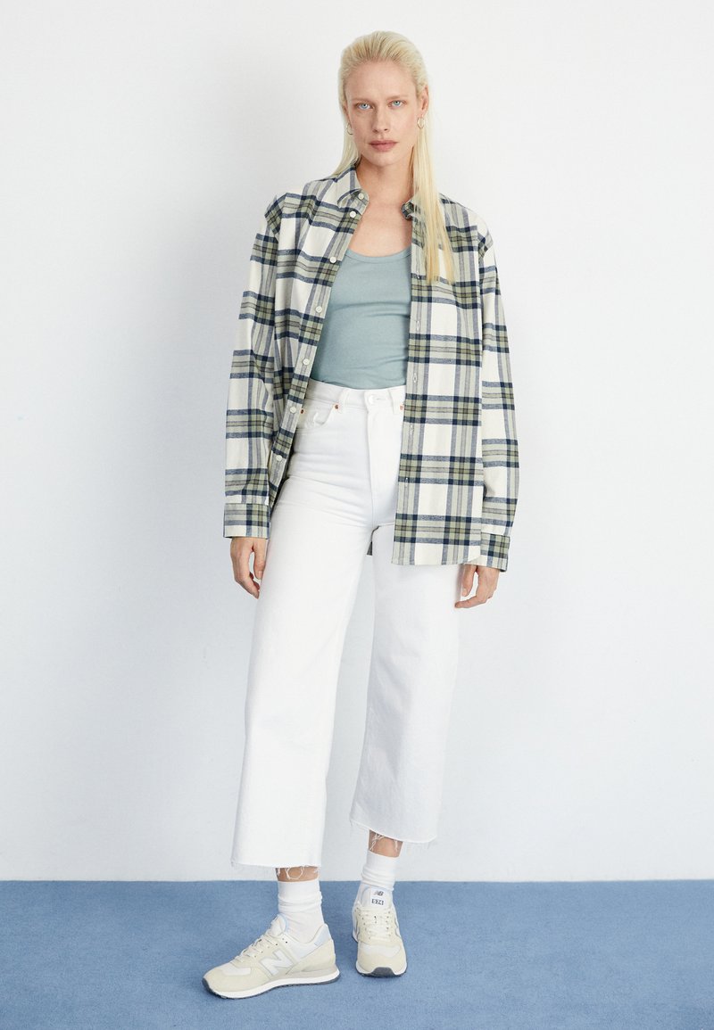Übergröße Karo-Flanellhemd in Creme, Blau und Grün; hellblaues Tanktop; weiße, cropped Jeans; beiges Sneakers mit weißen Akzenten.