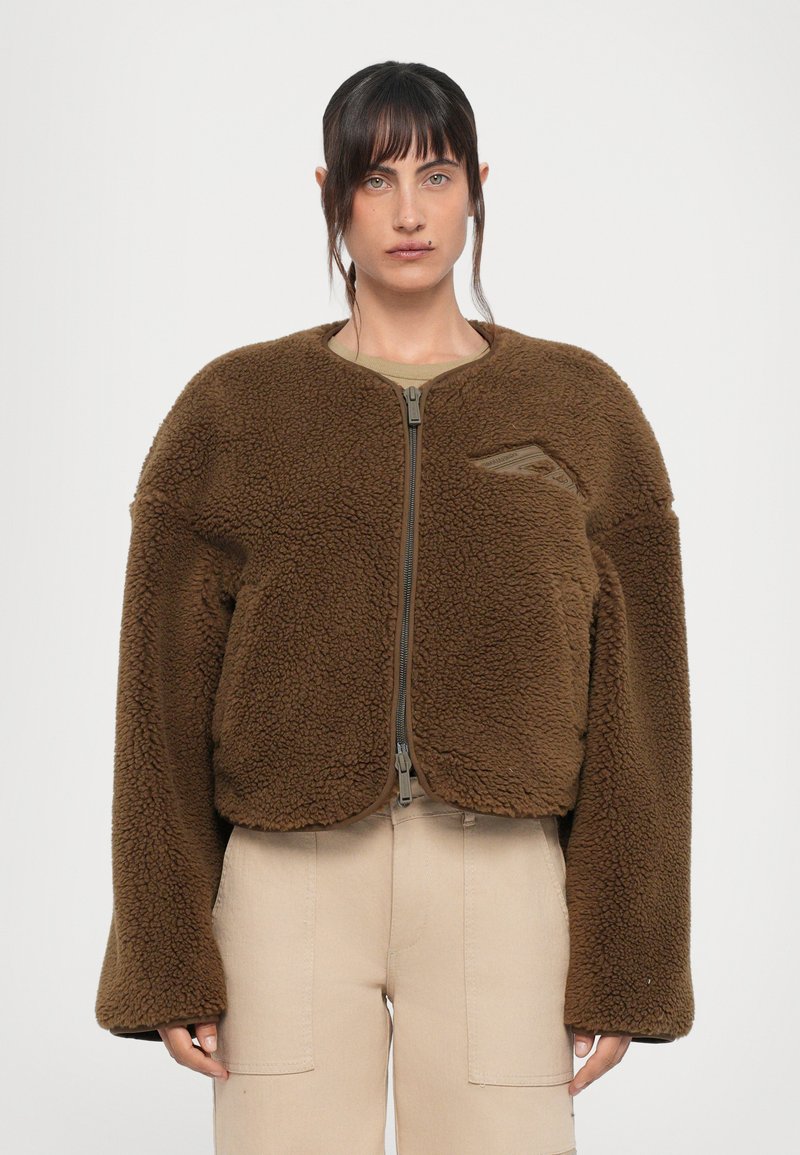 Braune Fleecejacke mit rundem Ausschnitt, Reißverschluss und überschnittenen Schultern. Sie verfügt über eine kleine Brusttasche auf der linken Seite und hat eine weiche Textur.