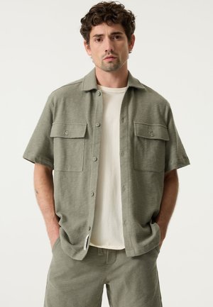 Lichtgroen shirt met korte mouwen, gemaakt van gestructureerde stof, met twee borstzakken en knoopsluiting, gedragen over een wit T-shirt.