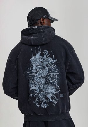 Persona que lleva una sudadera negra con capucha con un gran diseño detallado de un dragón plateado y floral en la espalda, combinada con una gorra negra de malla.