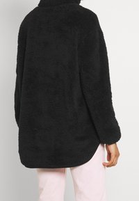 Schwarze, flauschige, übergroße Fleecejacke mit hohem Kragen, langen Ärmeln und abgerundetem Saum, die eine weiche Textur und ein lässiges Design aufweist.