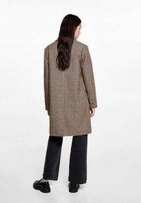 Dolga, karirasta plašč v rjavih, rdečih in kremnih odtenkih. Ima sproščen kroj, dolge rokave in srednje dolžino. V kombinaciji z črnimi širokimi hlačami.