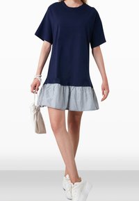 Navy T-Shirt-Kleid mit kurzen Ärmeln, gestreiftem Rüschensaum und lockerer Passform. Kombiniert mit einer weißen Tasche und weißen Sneakers.