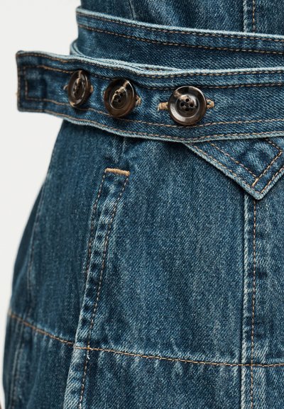Vêtement en denim avec une sangle réglable boutonnée comportant trois boutons foncés et des coutures orange visibles.