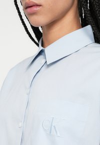 Gaiši zils krekls ar apkakli un kabatu, kurā ir balta izšūta "CK" logo. Gluda tekstūra un aizdare ar pogām.