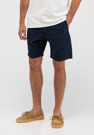 Man die marineblauwe korte broek, wit T-shirt en beige bootsschoenen draagt, staat met één hand in de zak tegen een effen achtergrond.
