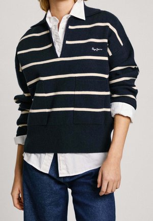 Person wearing navy blue and white striped sweater with front pockets over a white collared shirt and blue jeans.